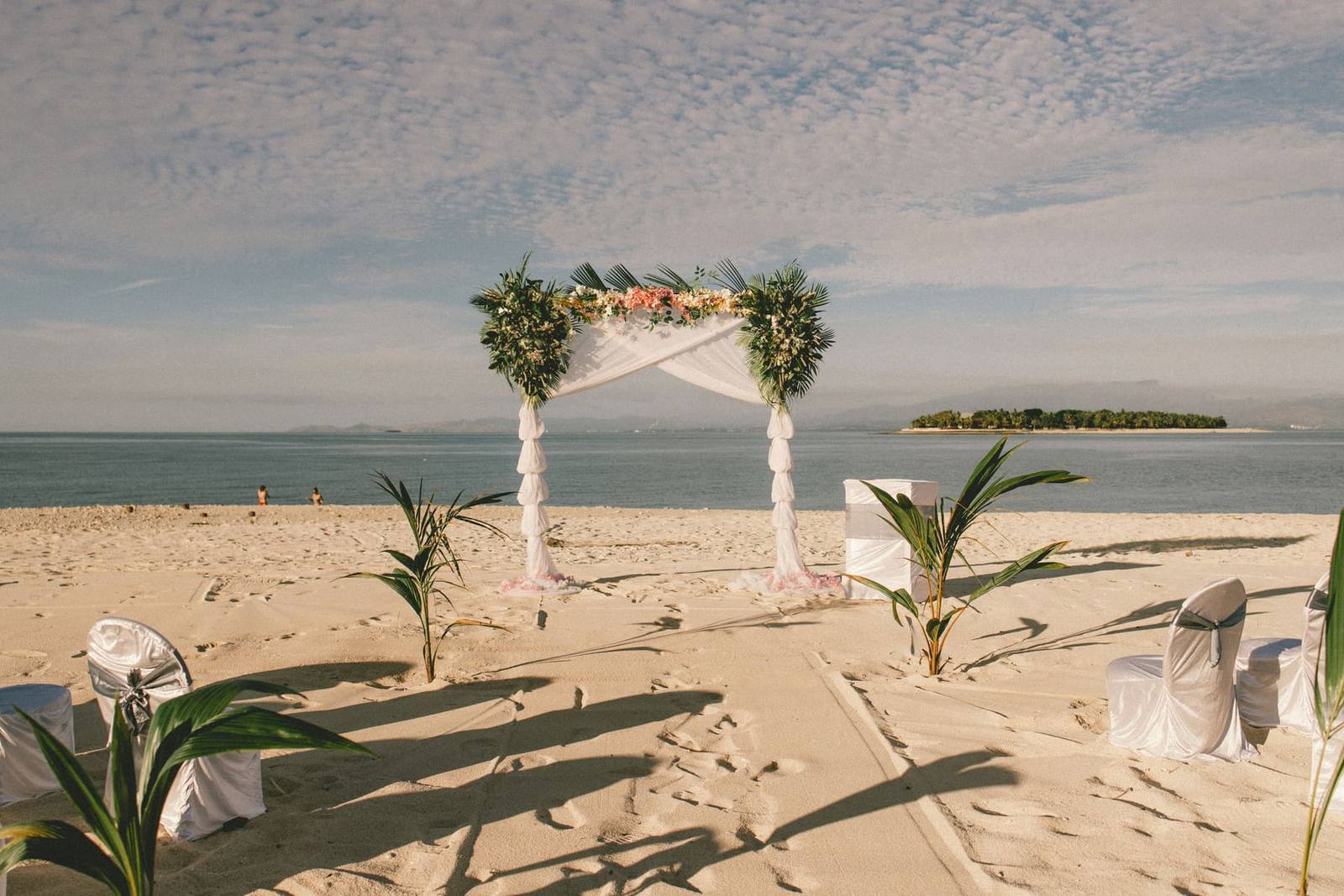 A floral wedding arch and white-draped chairs set up on a sandy beach beside calm water