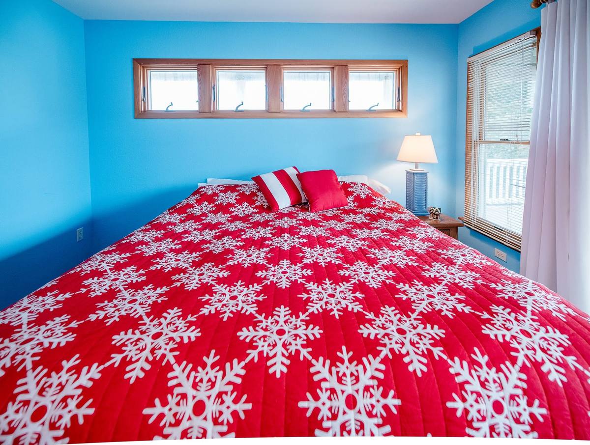 Close angle on a bed dressed in a red-and-white snowflake quilt beside a lit lamp and window in a blue bedroom