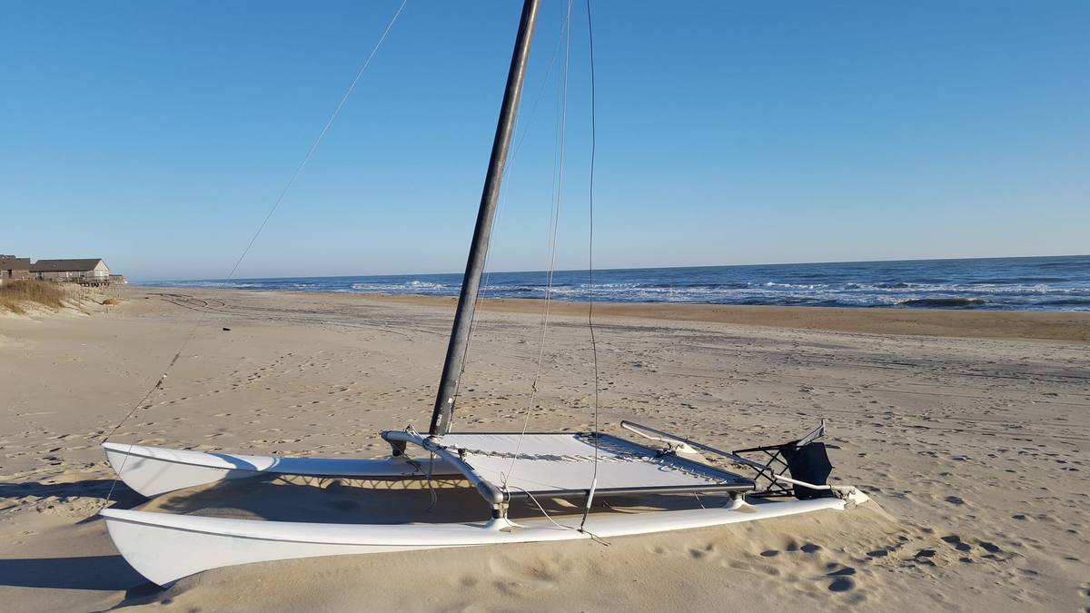 A small catamaran sailboat resting on the beach with the ocean behind it