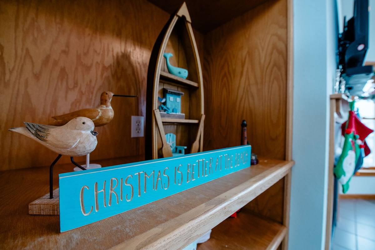 A teal wood sign reading Christmas is better at the beach displayed on built-in shelving alongside carved shorebird figures and a model sailboat