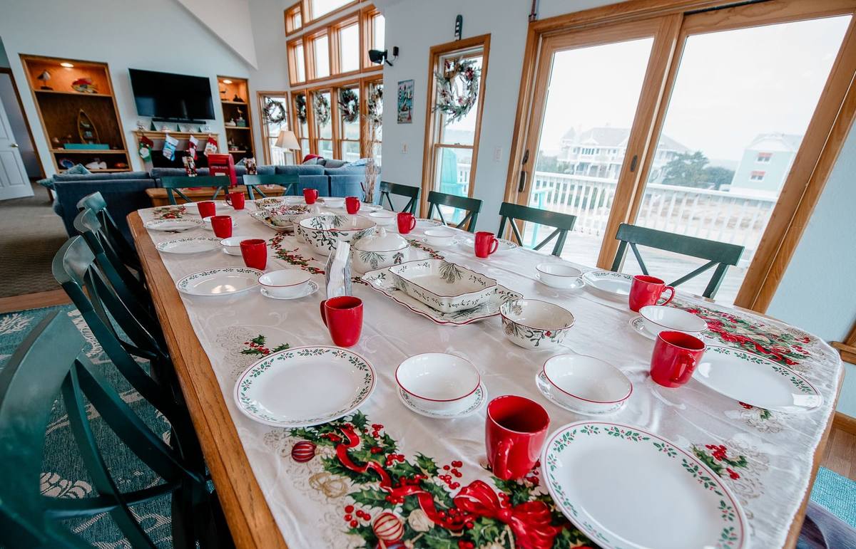 Close-up of a holiday-set dining table with a poinsettia table runner, red mugs, and holly-patterned place settings, wreaths visible in the windows beyond