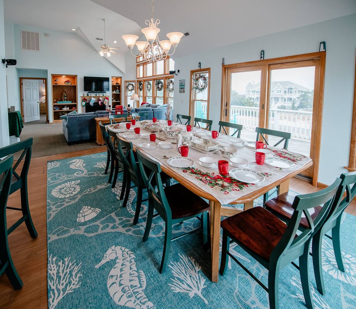 Long dining table set with a holiday runner and red mugs, seen across an open floor plan with wreath-hung windows, the mantle, and an ocean-town view beyond