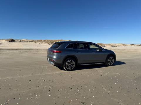 Electric SUV parked on the open sand beside dune grass, facing the ocean under a clear blue sky