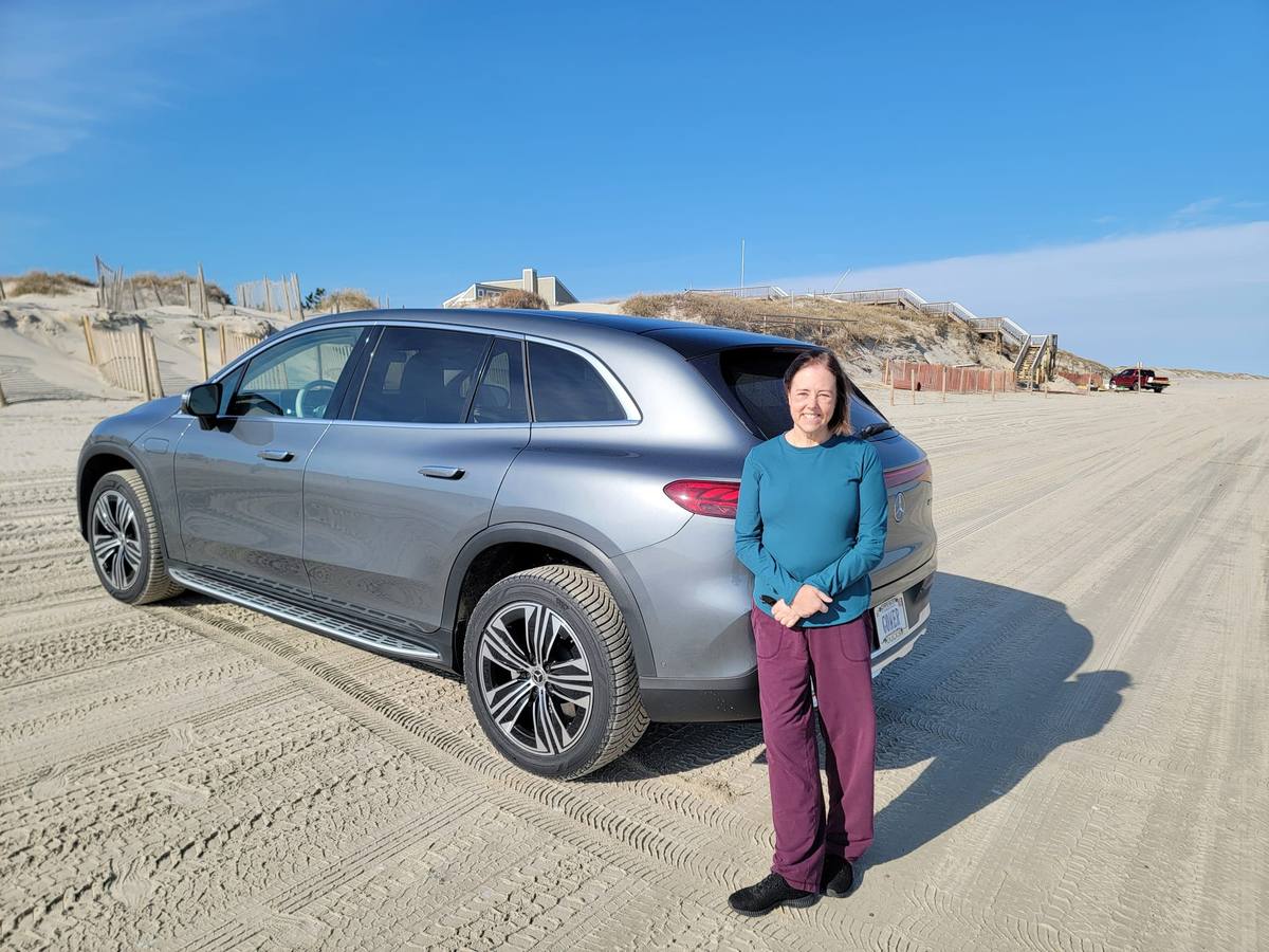 A woman standing beside a silver electric SUV parked on the open beach