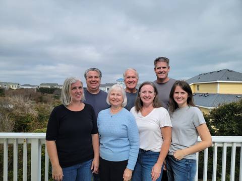 A seven-person multi-generational family posing together on a deck overlooking the dunes