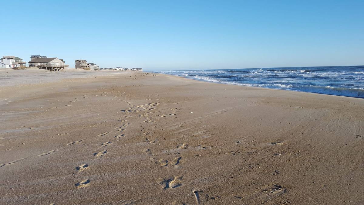 Footprints across a wide, empty beach leading toward the surf line with houses in the distance