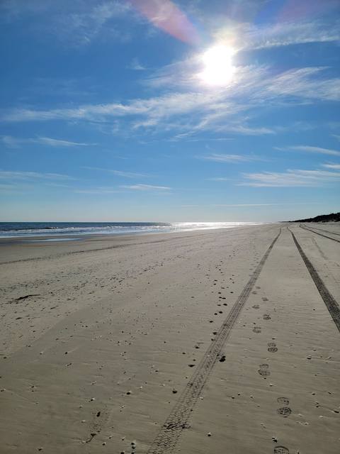 Vehicle tracks running along an empty beach beside the ocean under a blue sky with wispy clouds