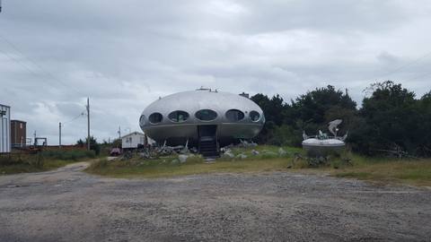 A round, saucer-shaped Futuro house with porthole windows sitting in a gravel lot