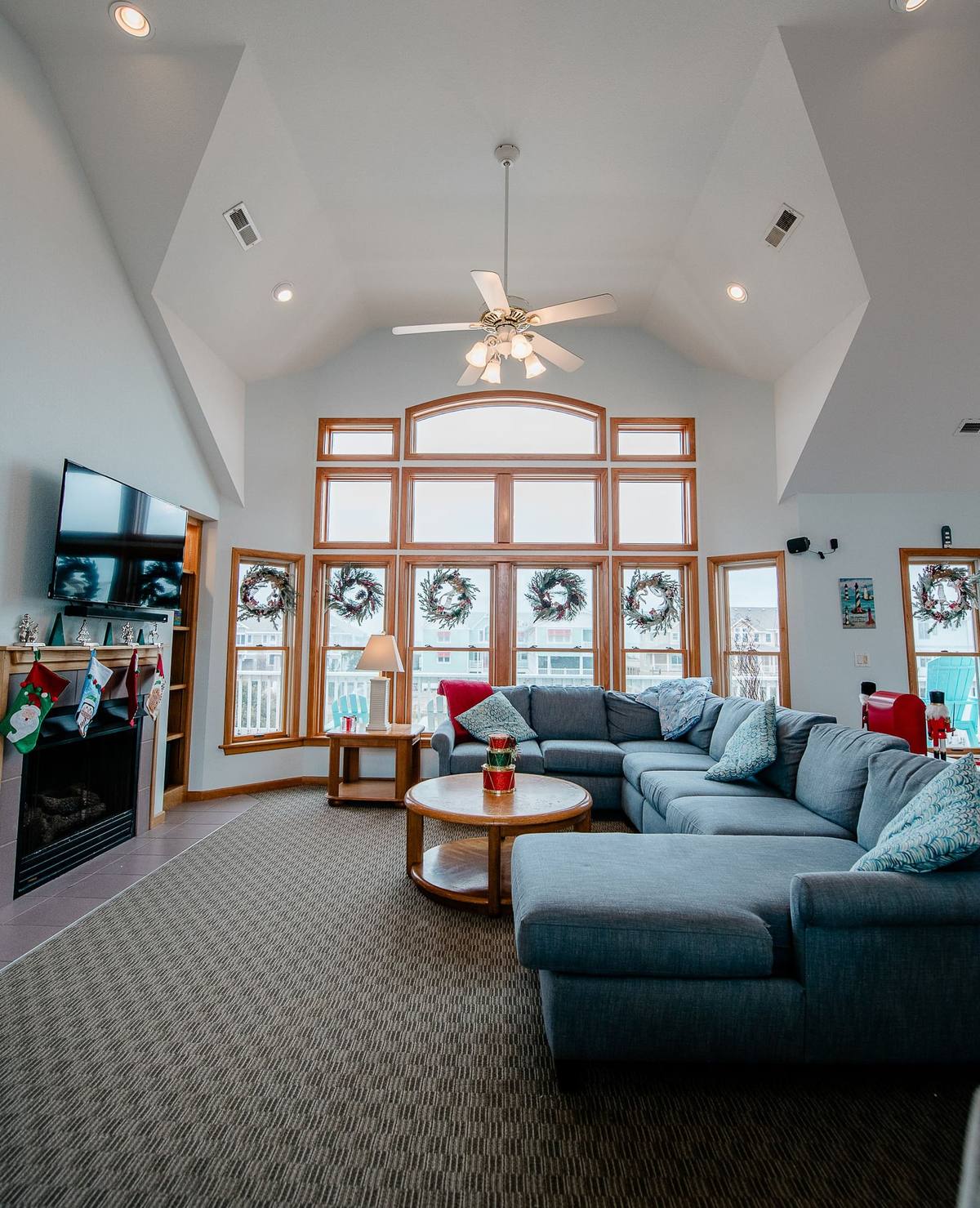 Vaulted great room with a wall of wreaths hung in the windows, a stocking-draped mantle to the left, and a sectional sofa facing the ocean-view windows