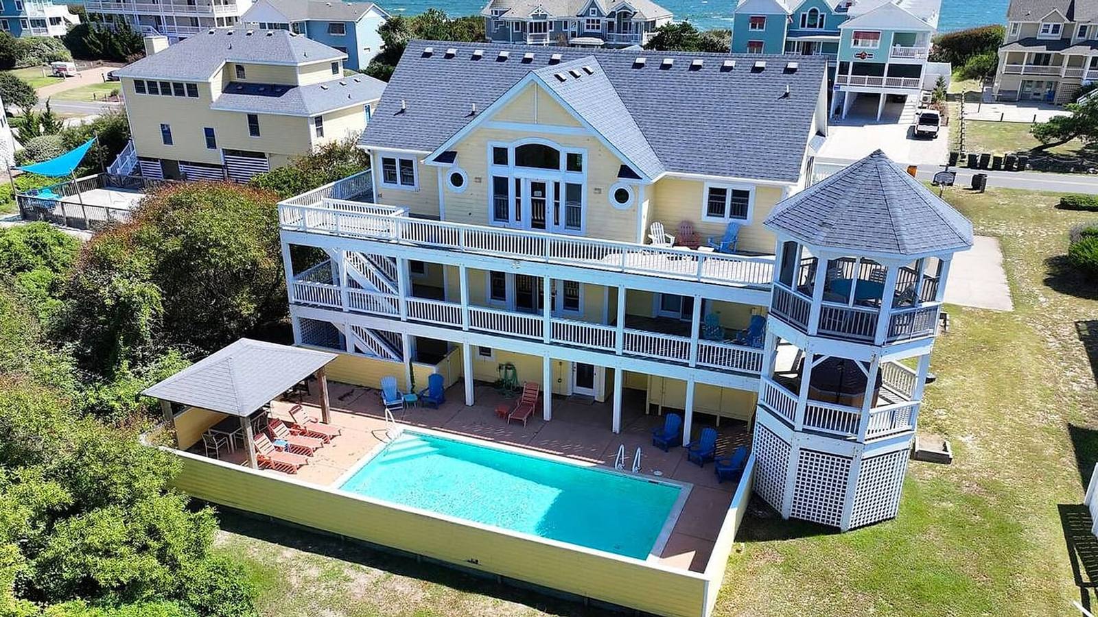 Aerial view of OBX Beach Bliss, a yellow three-level house with wraparound decks, a gazebo tower, and a heated pool, with the ocean beyond
