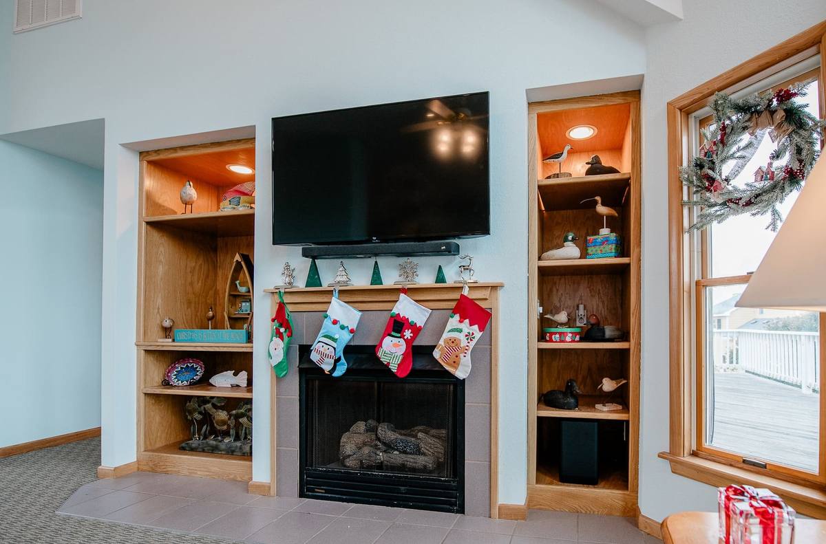 Wood mantle over the gas fireplace with four holiday stockings hung below a TV, flanked by built-in shelving and a wreath visible in the window