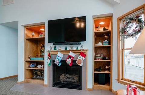 Wood mantle over the gas fireplace with four holiday stockings hung below a TV, flanked by built-in shelving and a wreath visible in the window