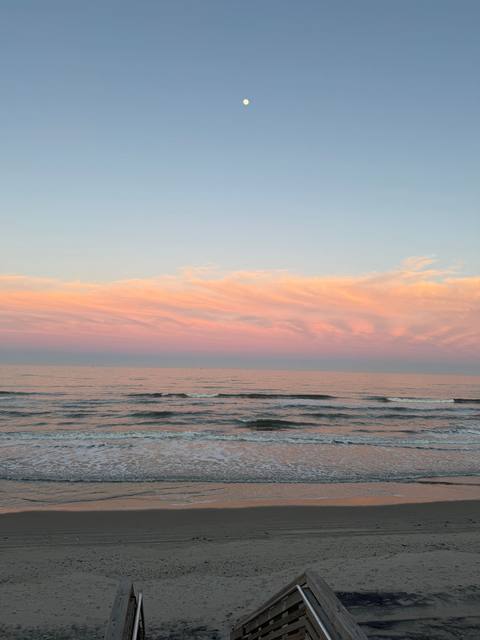 Pale pink sunset sky with a rising moon above gentle ocean waves, viewed from a wooden deck railing