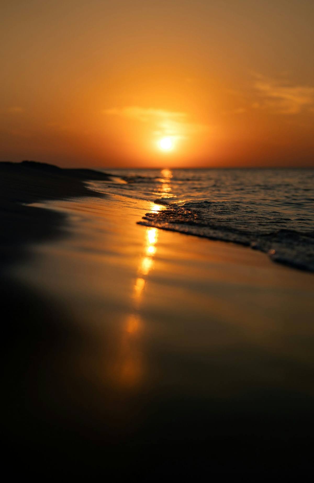 The sun low over a flat ocean horizon, glowing gold and reflecting off wet sand and gentle waves