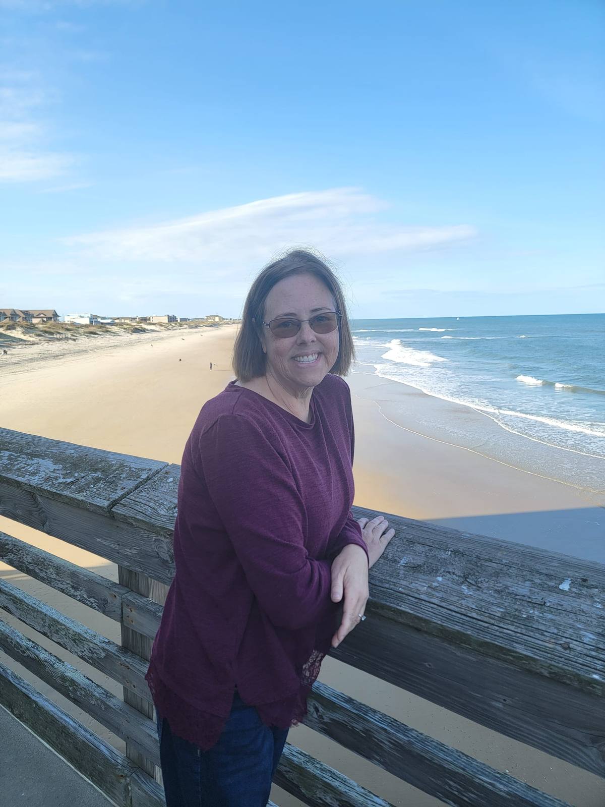 A woman smiling and leaning on a wooden pier railing overlooking the beach