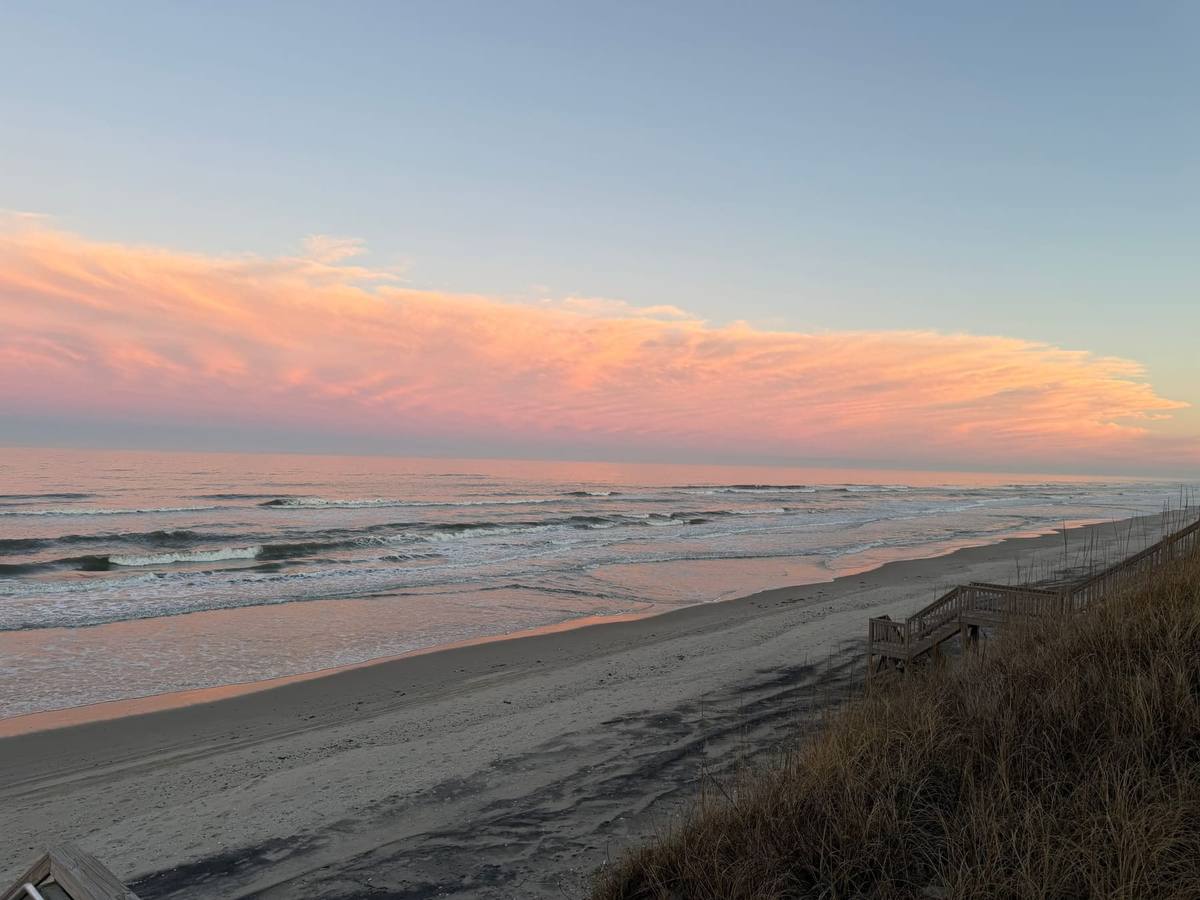 Pink and orange clouds stretching over the ocean at sunset, with the beach curving away below