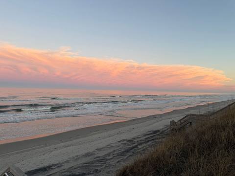 Pink and orange clouds stretching over the ocean at sunset, with the beach curving away below
