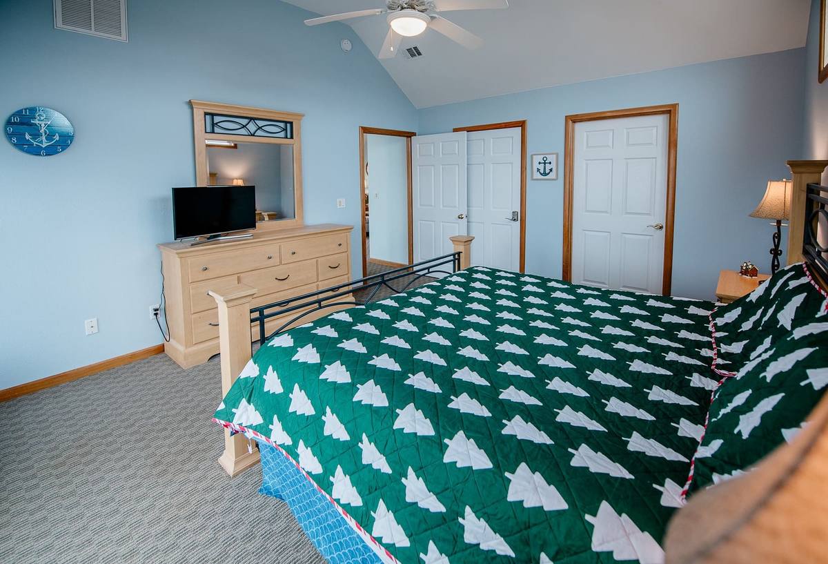 Light-blue primary bedroom with a vaulted ceiling and a king bed dressed in a dark green quilt patterned with white evergreen trees