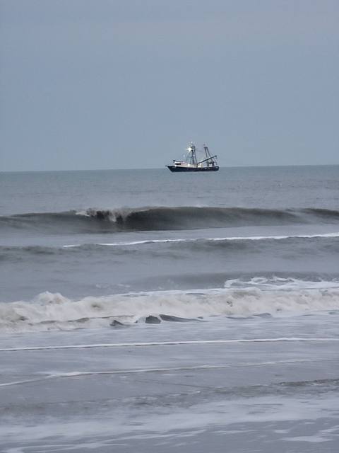 A commercial shrimp trawler on the horizon beyond breaking waves on a gray, overcast winter day