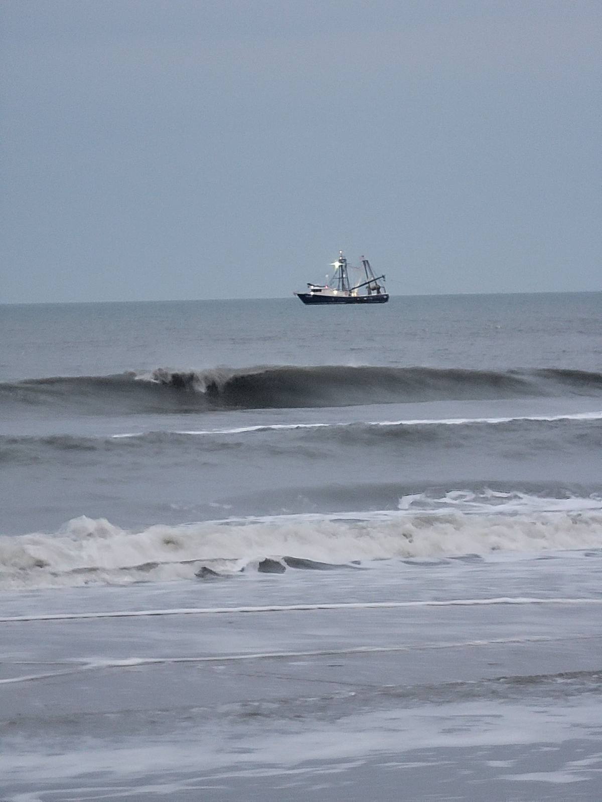 A commercial shrimp trawler on the horizon beyond breaking waves on a gray, overcast winter day
