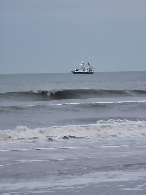 A shrimp trawler on the horizon beyond breaking waves under a gray sky