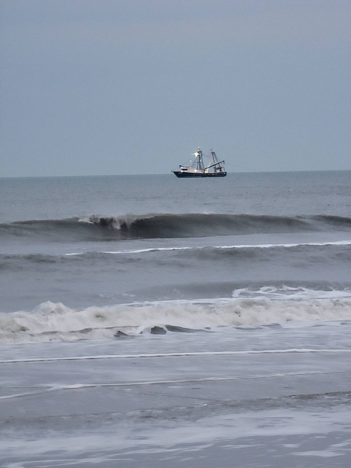 A shrimp trawler on the horizon beyond breaking waves under a gray sky