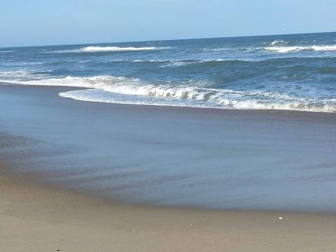 Small waves rolling up a wide, wet stretch of Outer Banks beach under a blue sky