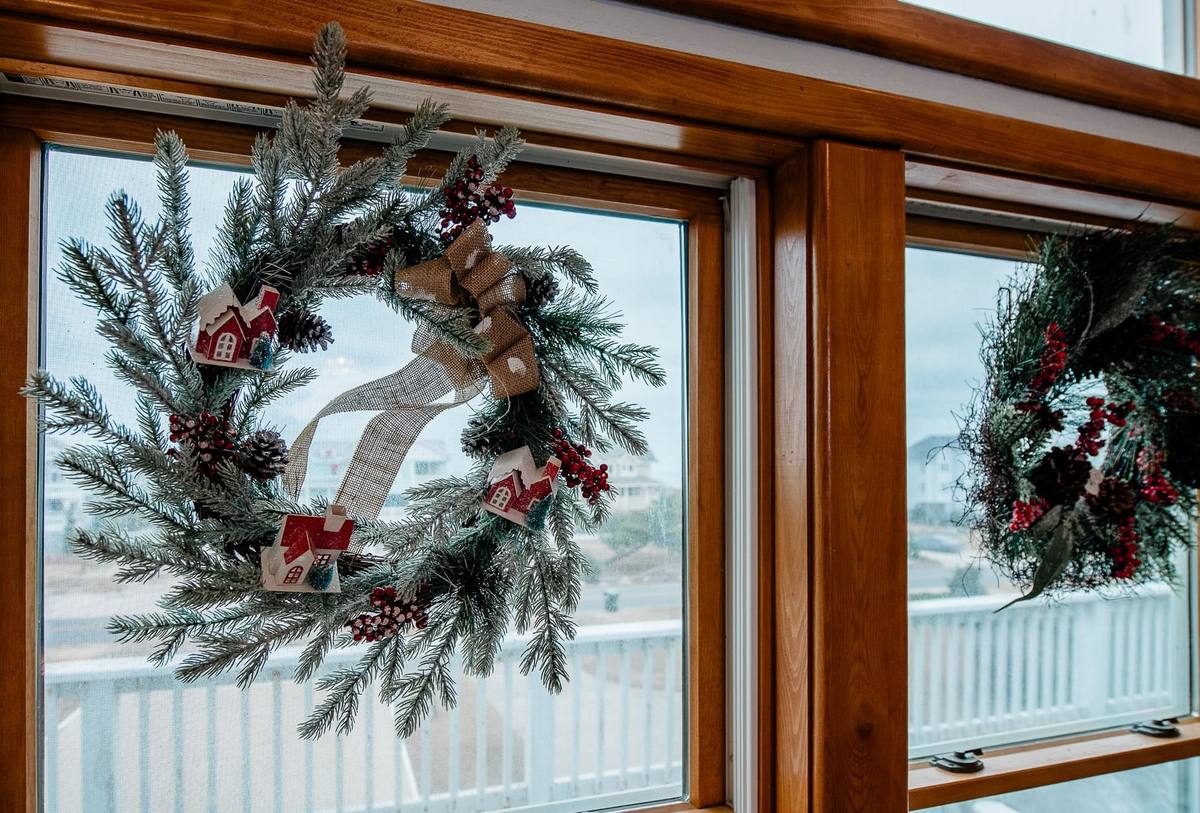 A frosted pinecone wreath with a burlap bow hangs in a wood-framed window, with the beach community and railing visible outside