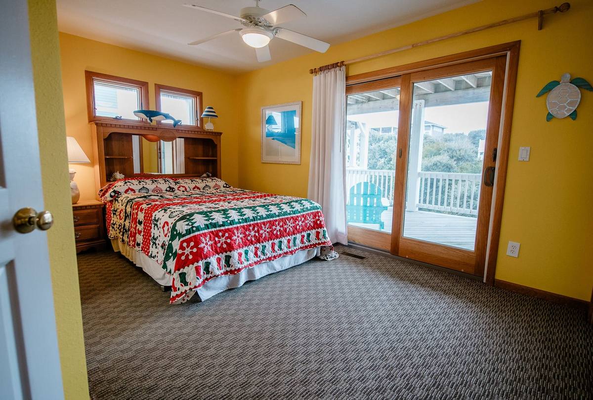 Yellow bedroom with a red-and-green patterned holiday quilt on the bed, facing glass doors with a deck and ocean-community view